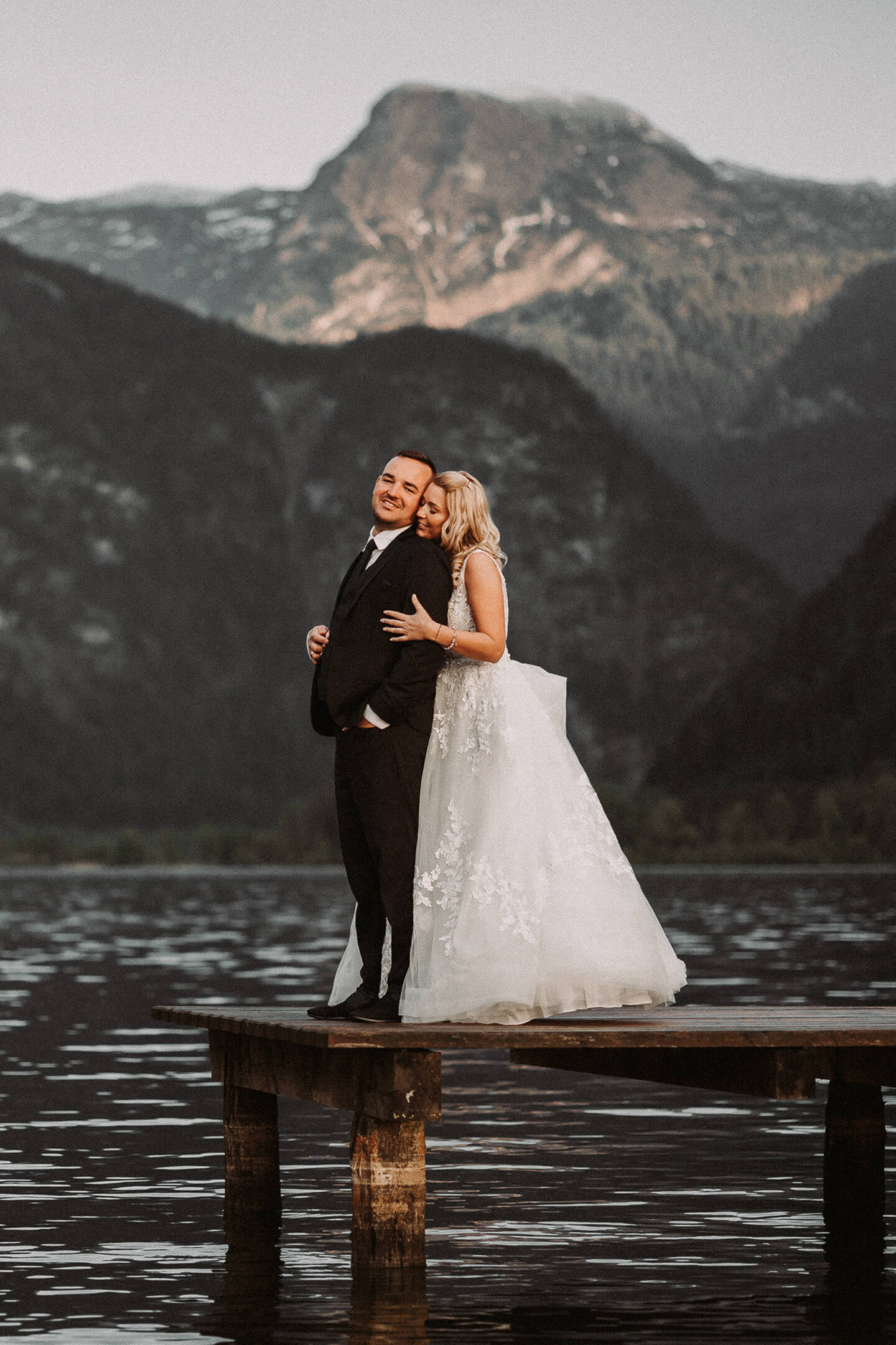A newlywed couple embraces each other while standing on a lakeside dock.