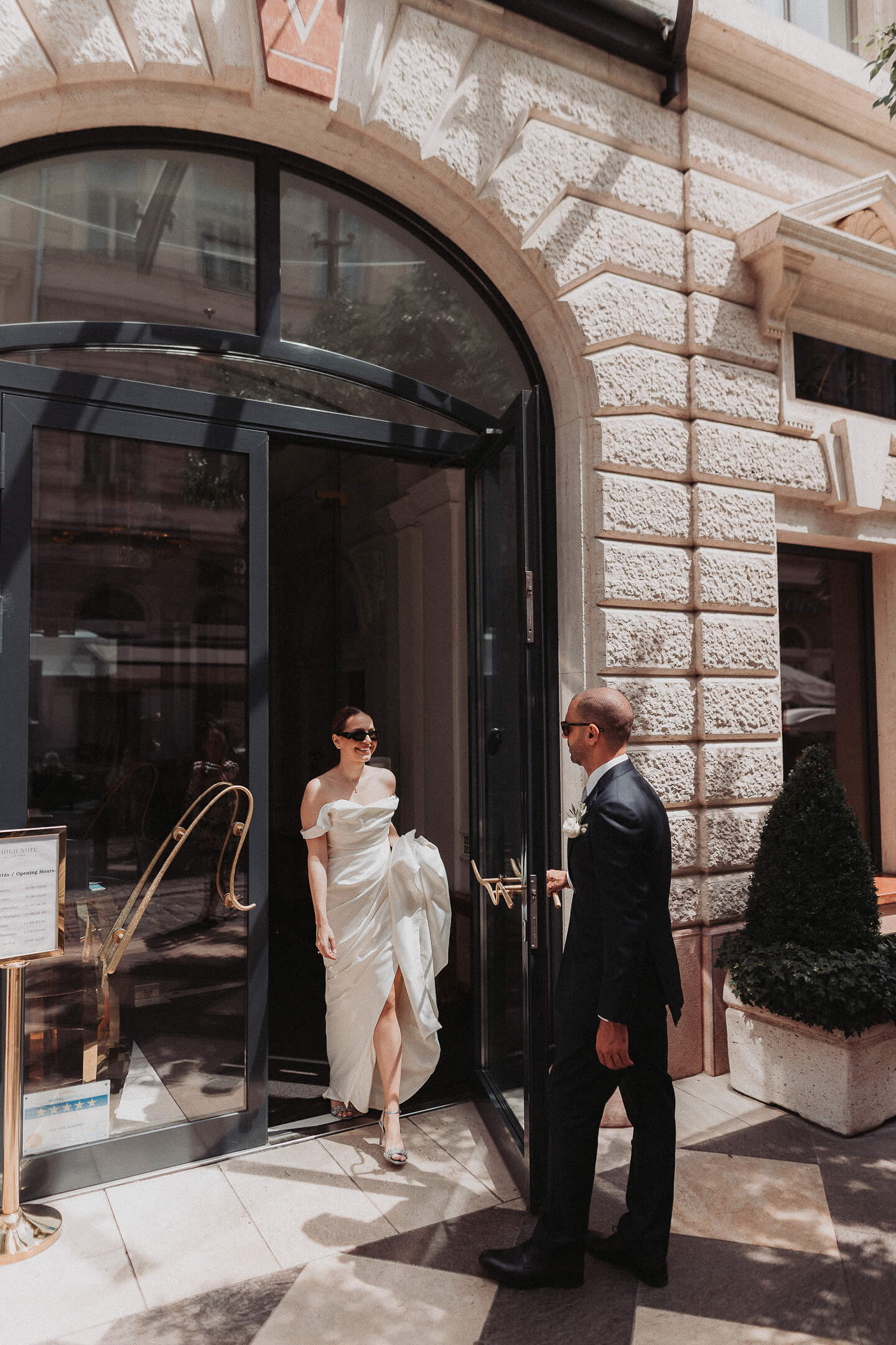 A bride steps out of an elegant building as the groom waits for her.