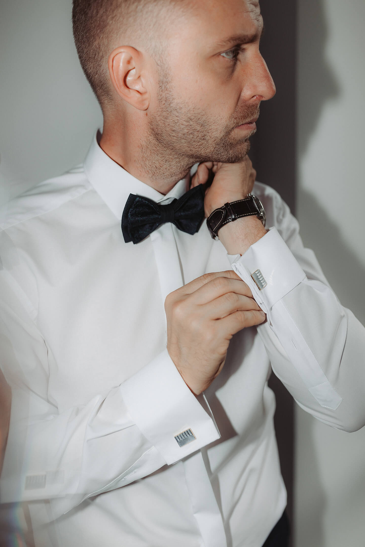 The groom adjusts his cufflinks, wearing an elegant white shirt and bow tie.