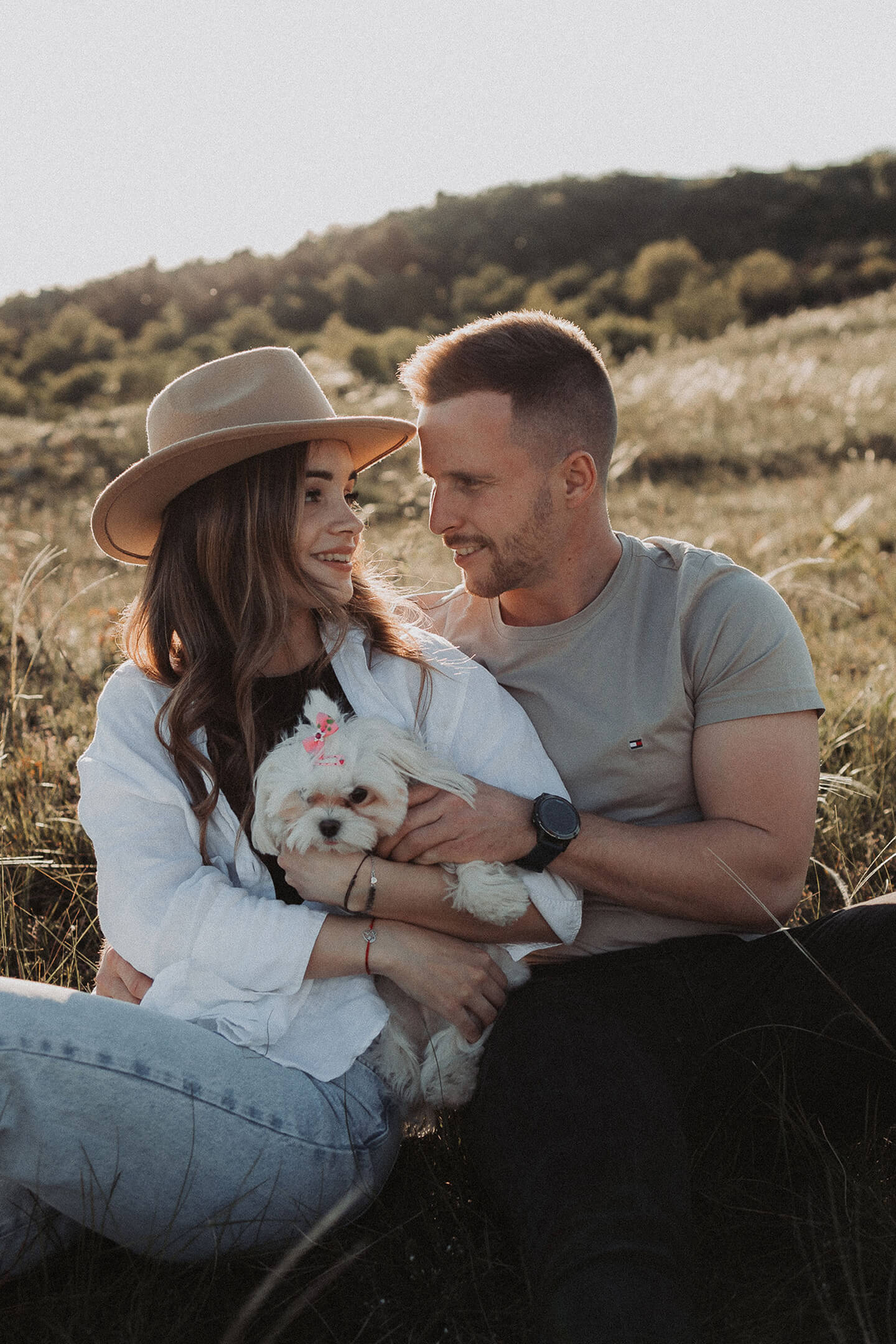 A young couple sits in a meadow with a small white dog, smiling at each other.